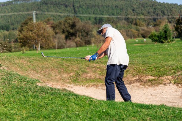 elderly golfer demonstrates skill and precision as he strikes the ball to lift it out of a sand bunker on the golf course. The focus is on the technique required to navigate the challenging sand trap