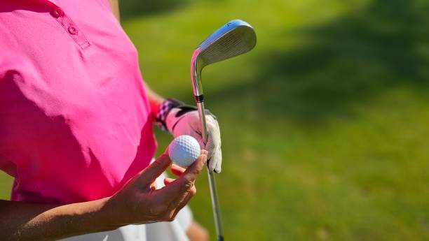 A golfer in a pink shirt prepares to tee off on a sunny day at a lush green course, holding a golf ball and club ready for the next shot