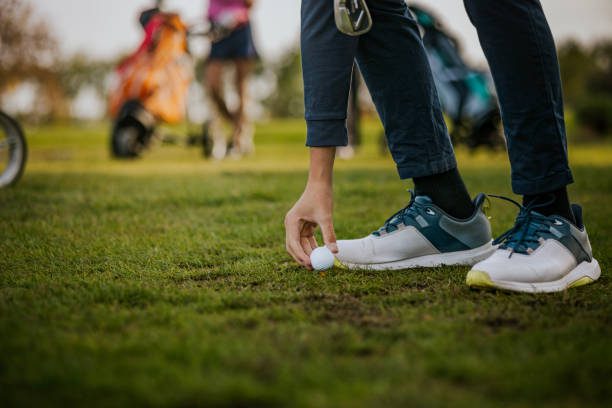 This image captures a golfer in the process of placing a golf ball on the tee, ready for a shot. The focus is on the player's hand and feet, showcasing the equipment and attire, with a golf course setting in the background. This scene is perfect for illustrating outdoor sports and leisure activities.