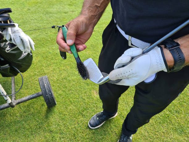 A golfer on a golf course is cleaning the grooves of their golf club with a green and black brush